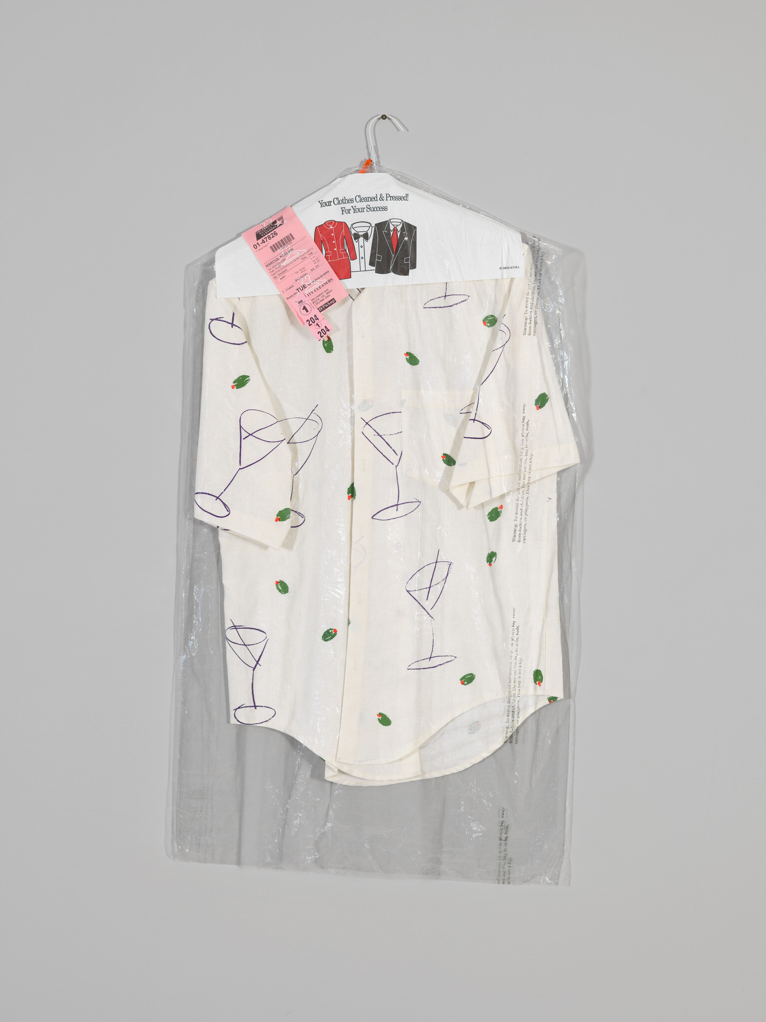 A white short-sleeve shirt with martini glass and olive print hangs in dry-cleaning plastic.