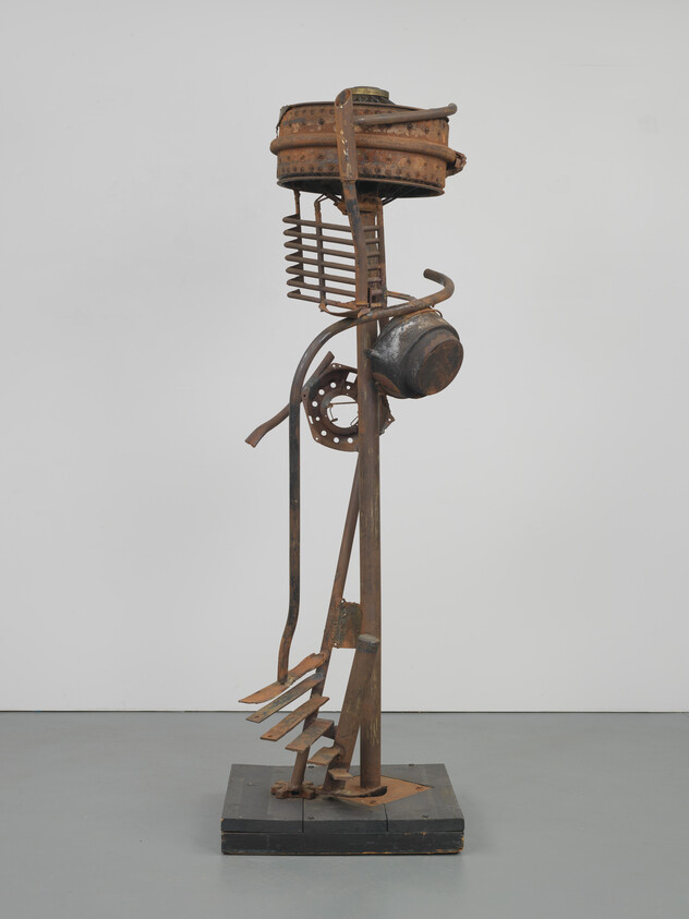 Tall vertical sculpture made from rusted scrap metal parts, including pots, gears, and curved rods.