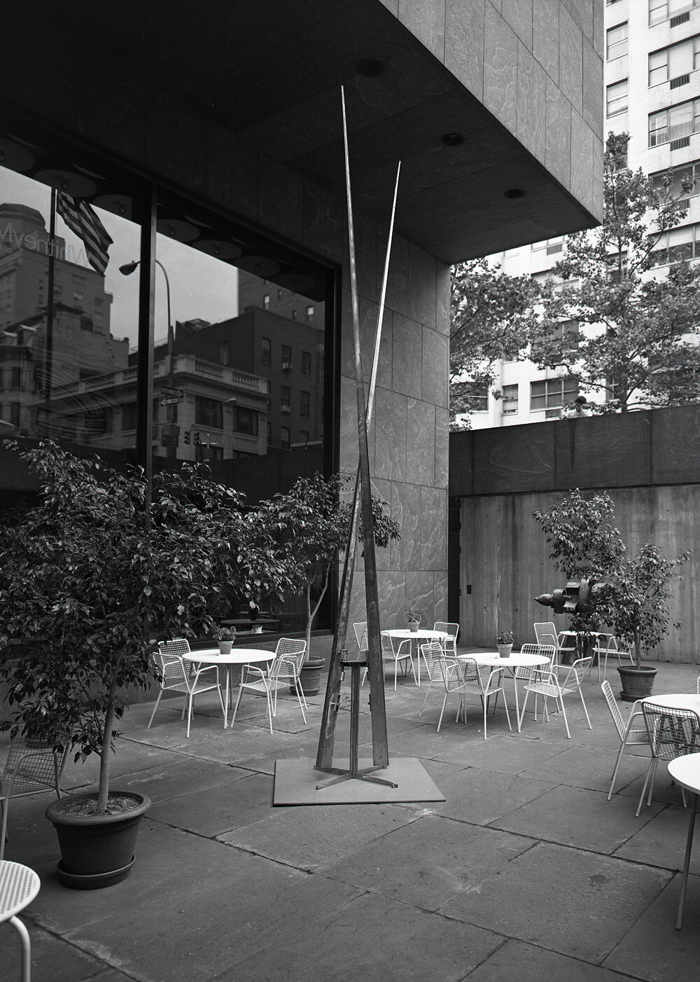 Tall slender metal sculpture stands in a courtyard surrounded by tables, chairs, and potted plants.