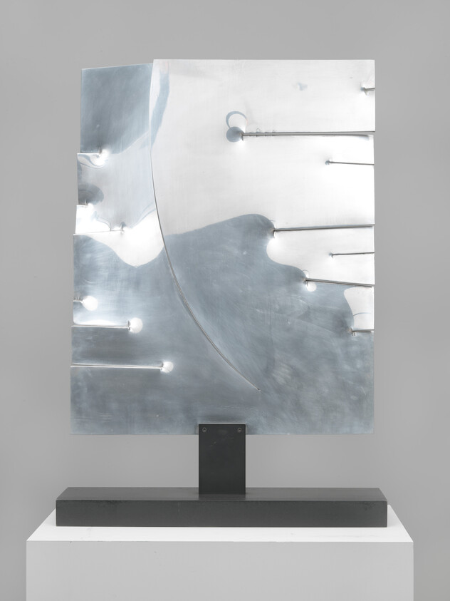 Abstract polished metal sculpture with curved cutouts and horizontal rods mounted on a black pedestal.