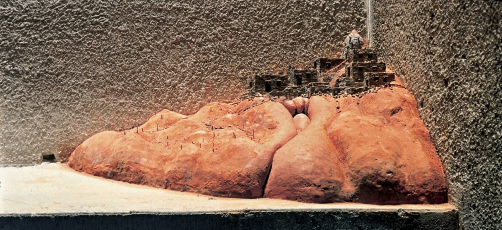 A small clay model of a reddish hill with miniature stone buildings and paths on top.