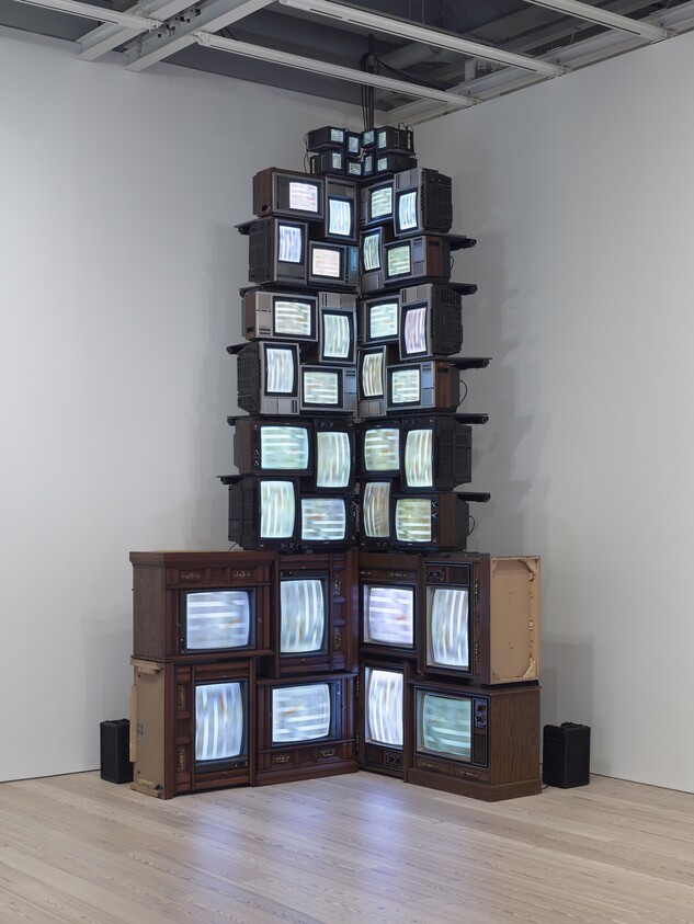 A tall stacked tower of vintage televisions in a gallery corner displaying moving static.