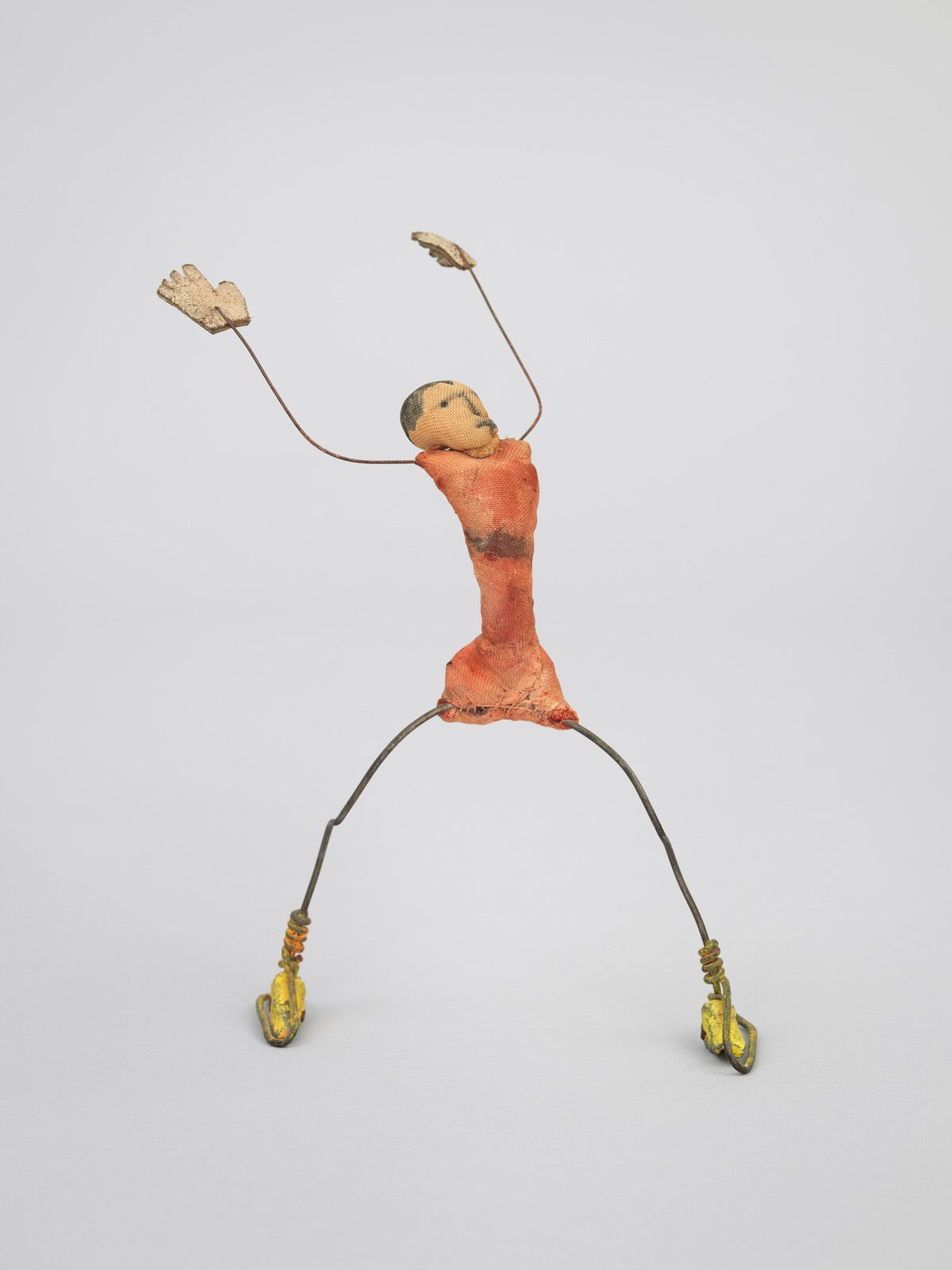A small wire sculpture of a person with raised arms, wearing an orange outfit and yellow shoes.