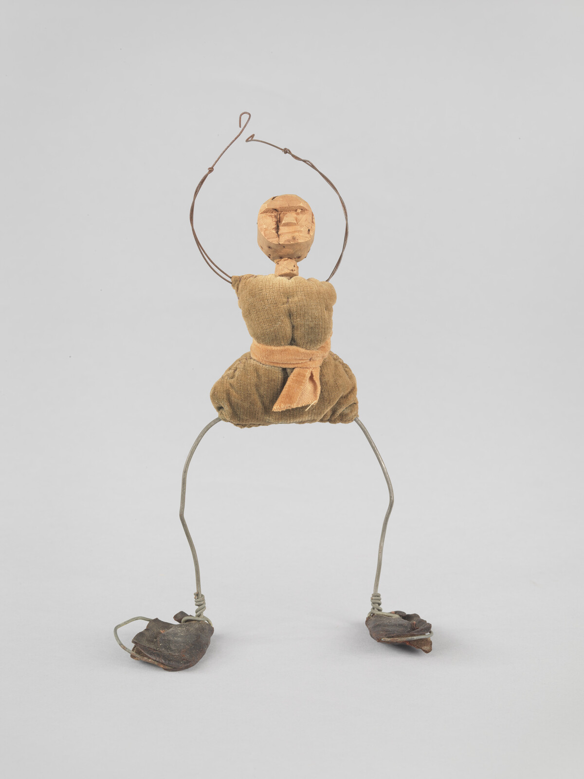 A simple wire and fabric figure with a wooden head, standing on a plain background.