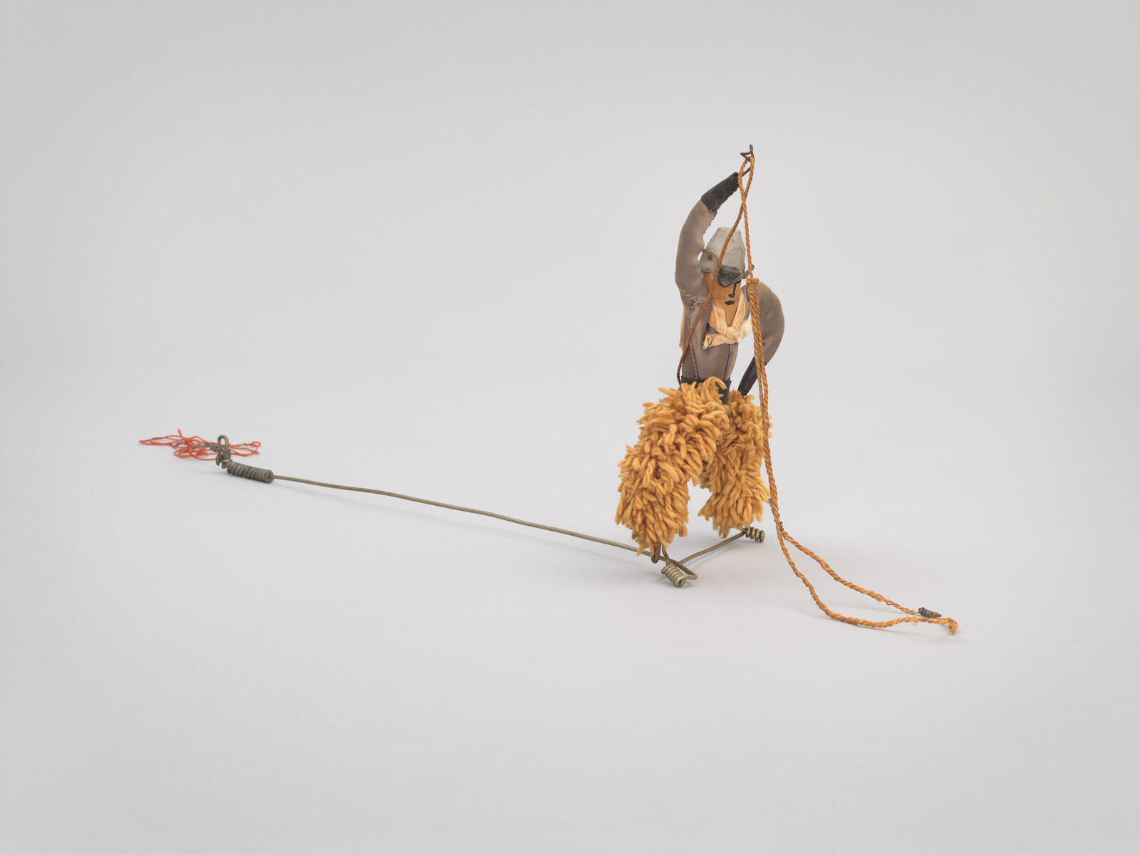 Small handmade figure with shaggy orange pants stands on a thin wire base holding an orange rope.