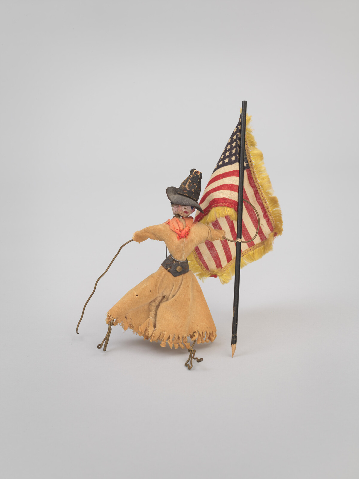 A small cloth doll dressed as a pioneer holds a fringed American flag while waving a wire arm.