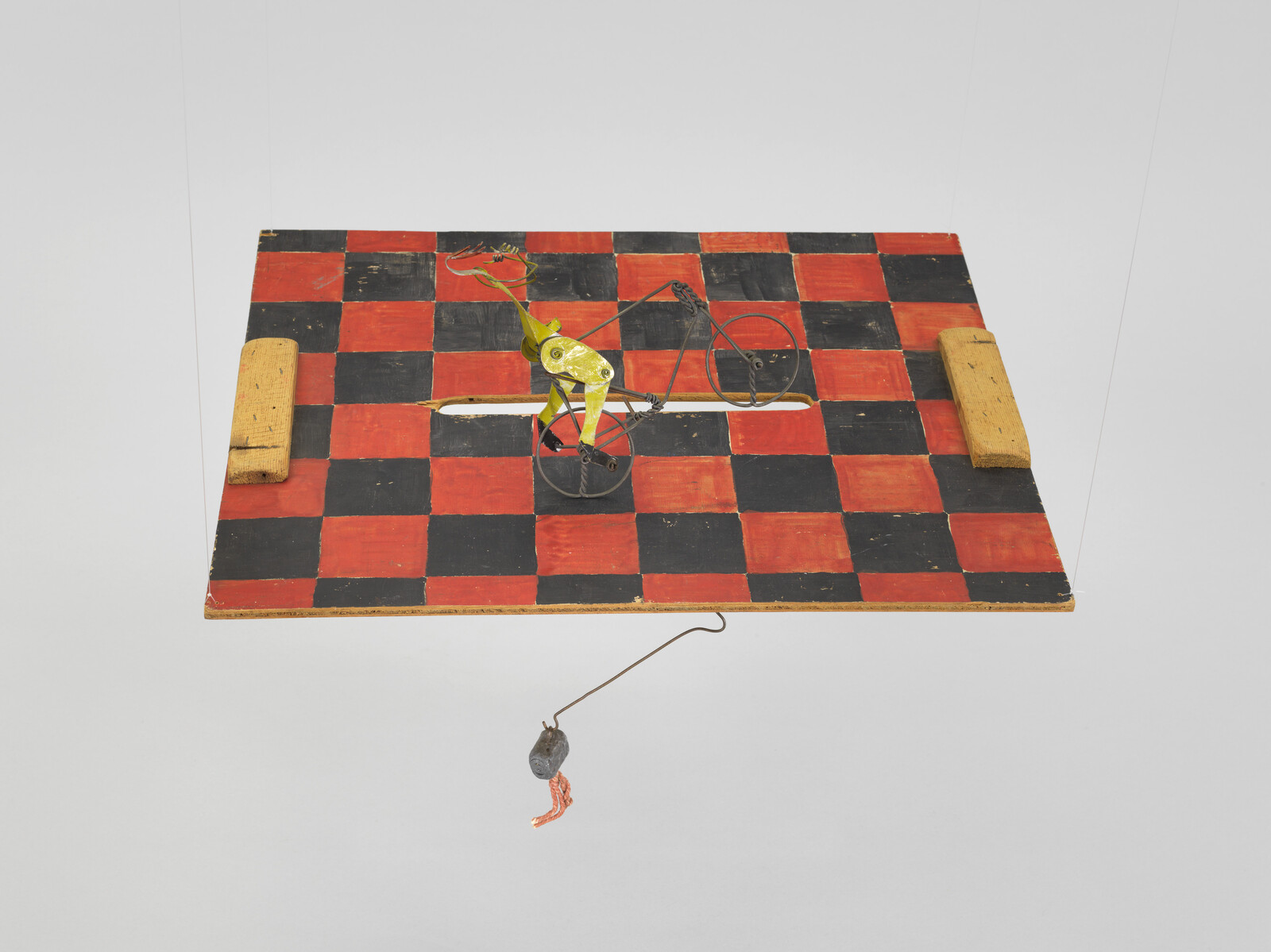 A yellow metal toy cyclist rides on a red and black checkered board with hanging weight.