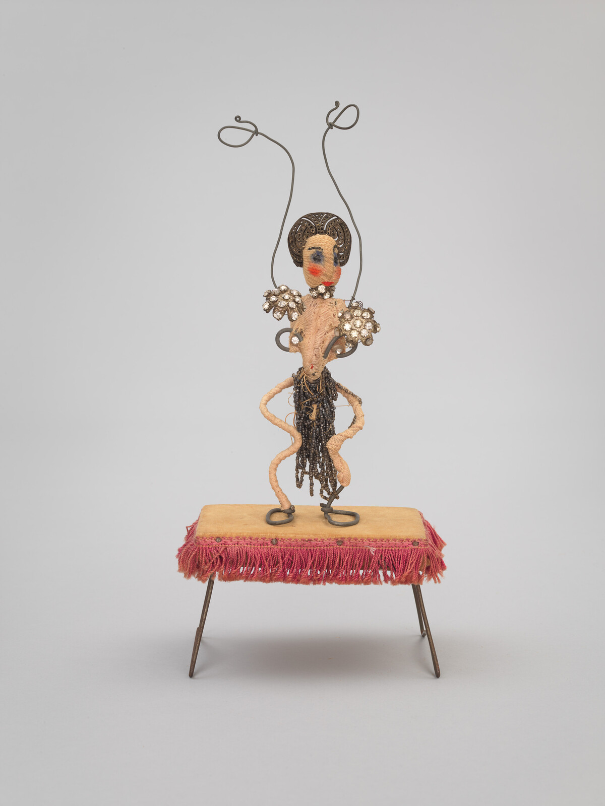 A small wire and bead figure with a decorated head stands on a fringed platform.