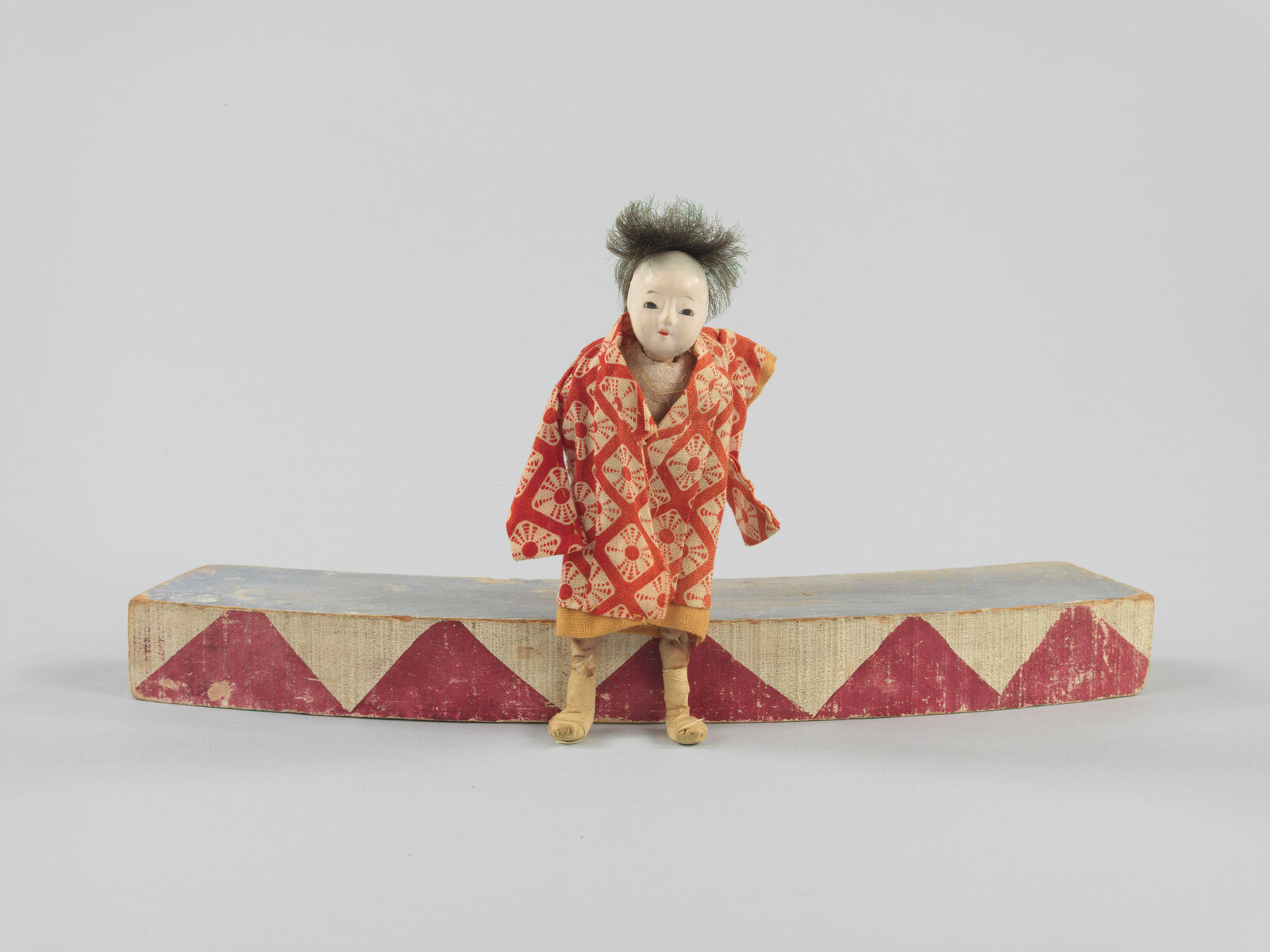 A small doll with a red patterned robe stands on a curved wooden platform with red triangles.