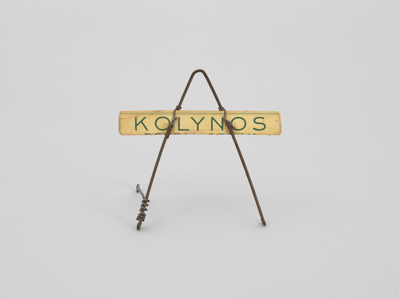 A small wire easel holds a rectangular label reading "KOLYNOS" elevated above a plain background.