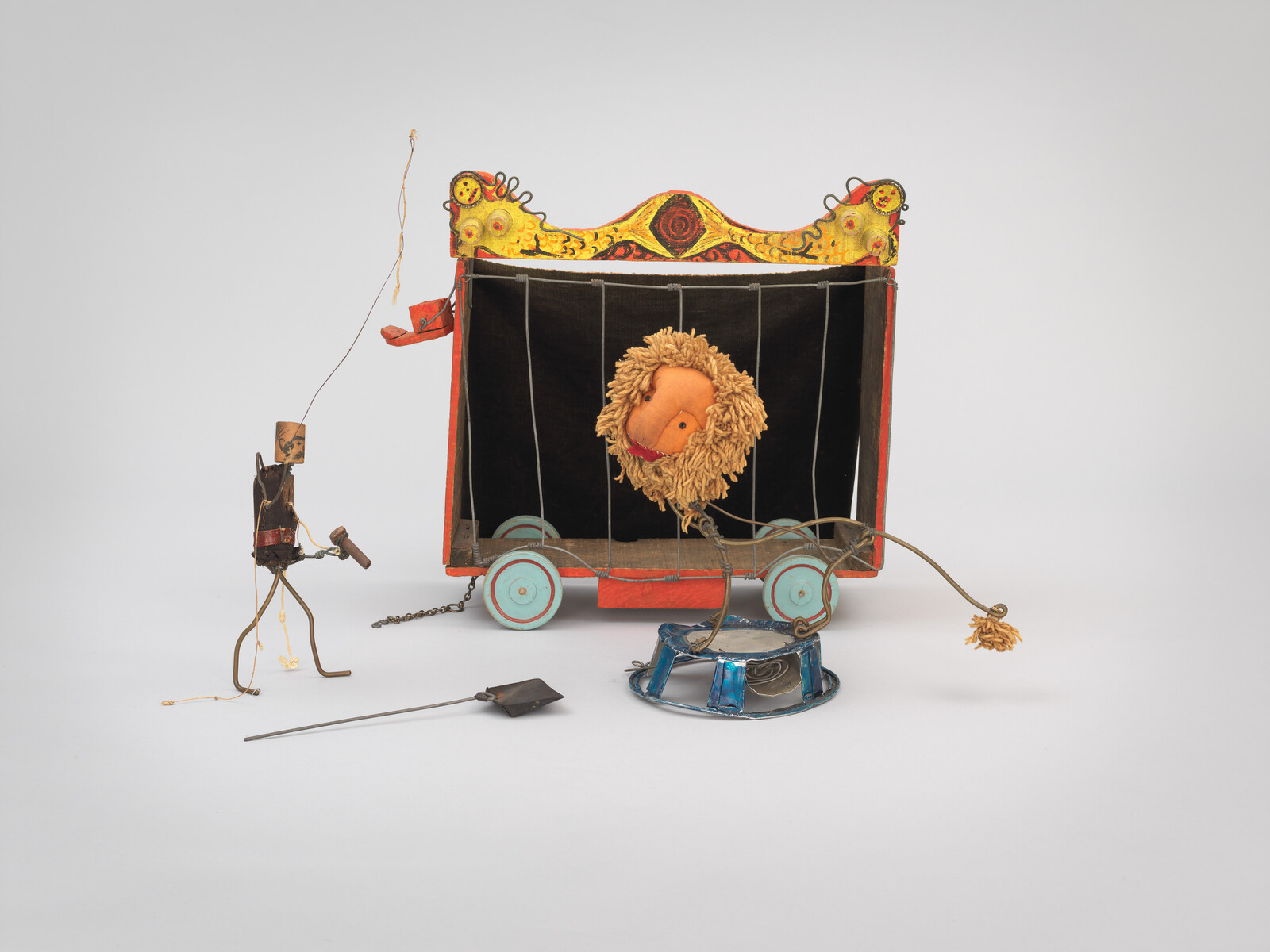 A toy lion puppet leans out of a small colorful wagon cage while a wire puppeteer stands nearby.