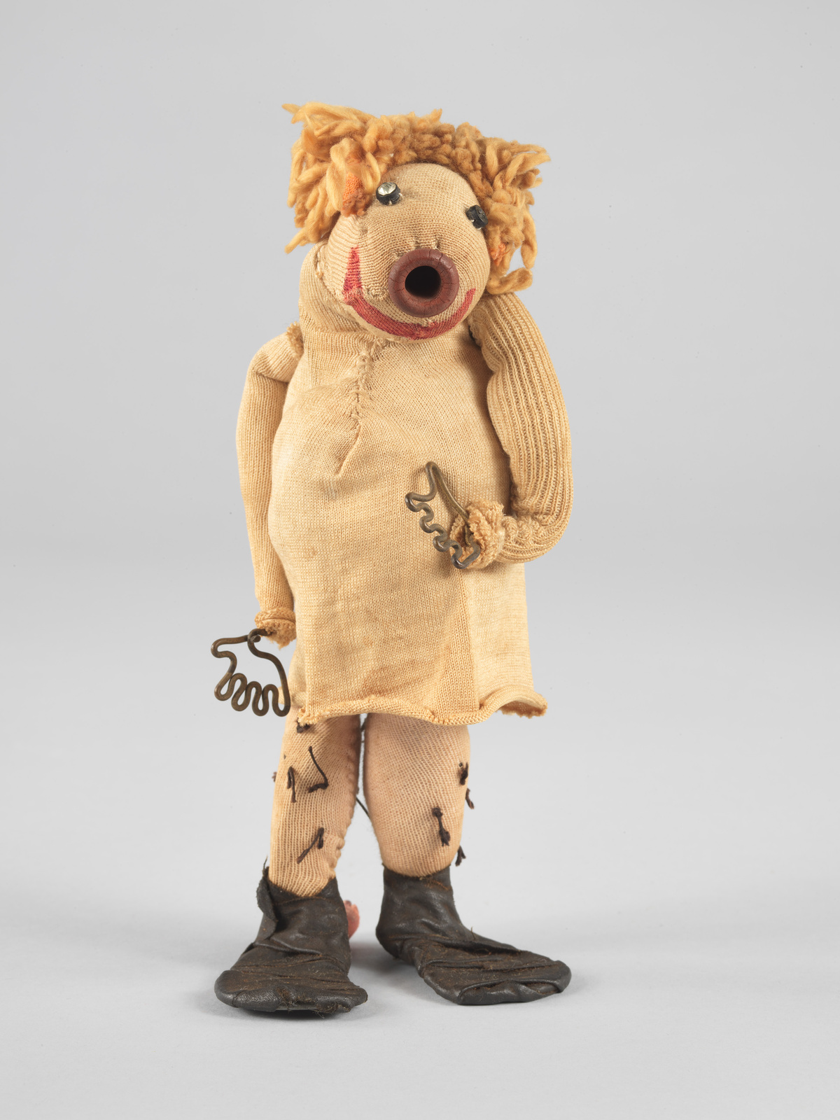 A rag doll-like sculpture, made of yellowed cloth with a round button-like nose, small rhinestone eyes, a curve for a smile, orange yarn hair, wire hands, and floppy shoes.