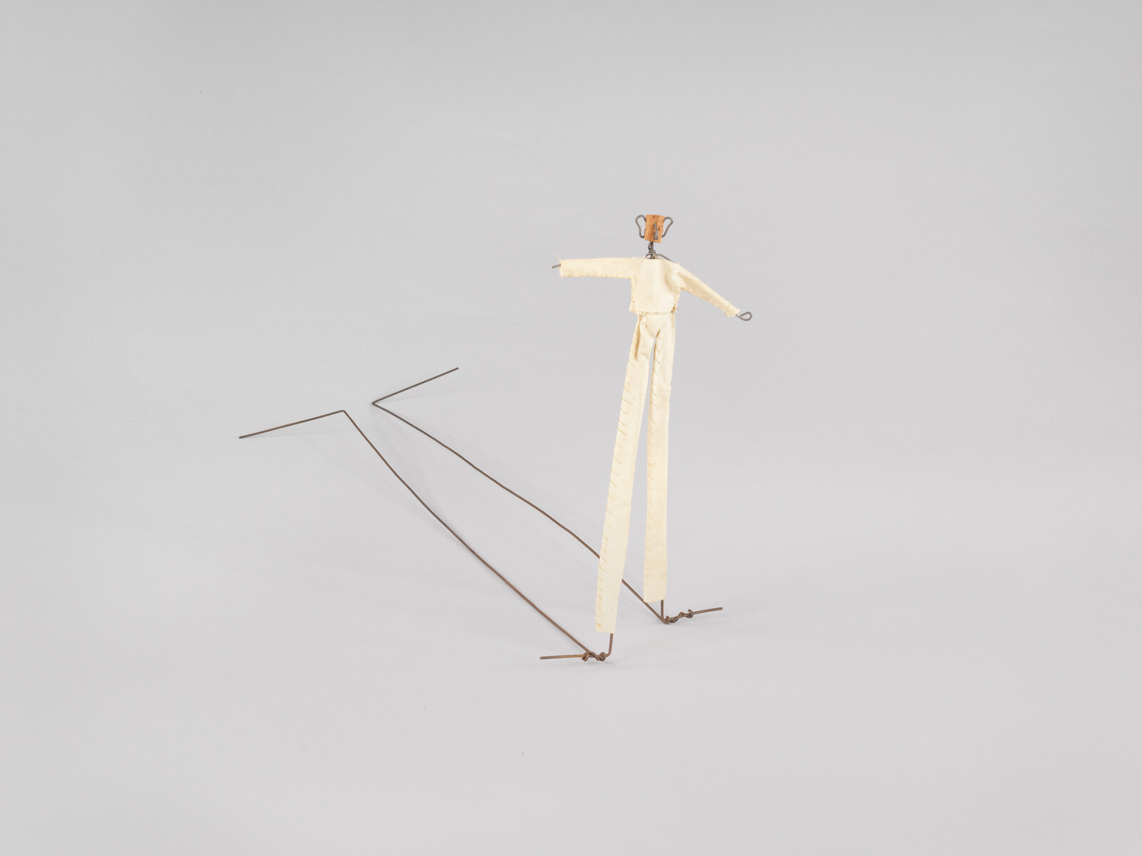 A small wire and fabric figure stands on tall wire stilts casting a long angular shadow.