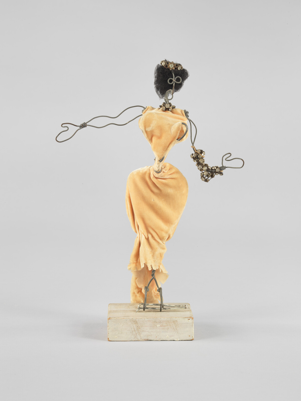 Small wire mannequin wearing a gathered yellow fabric dress and beaded bracelet, standing on a wooden block.