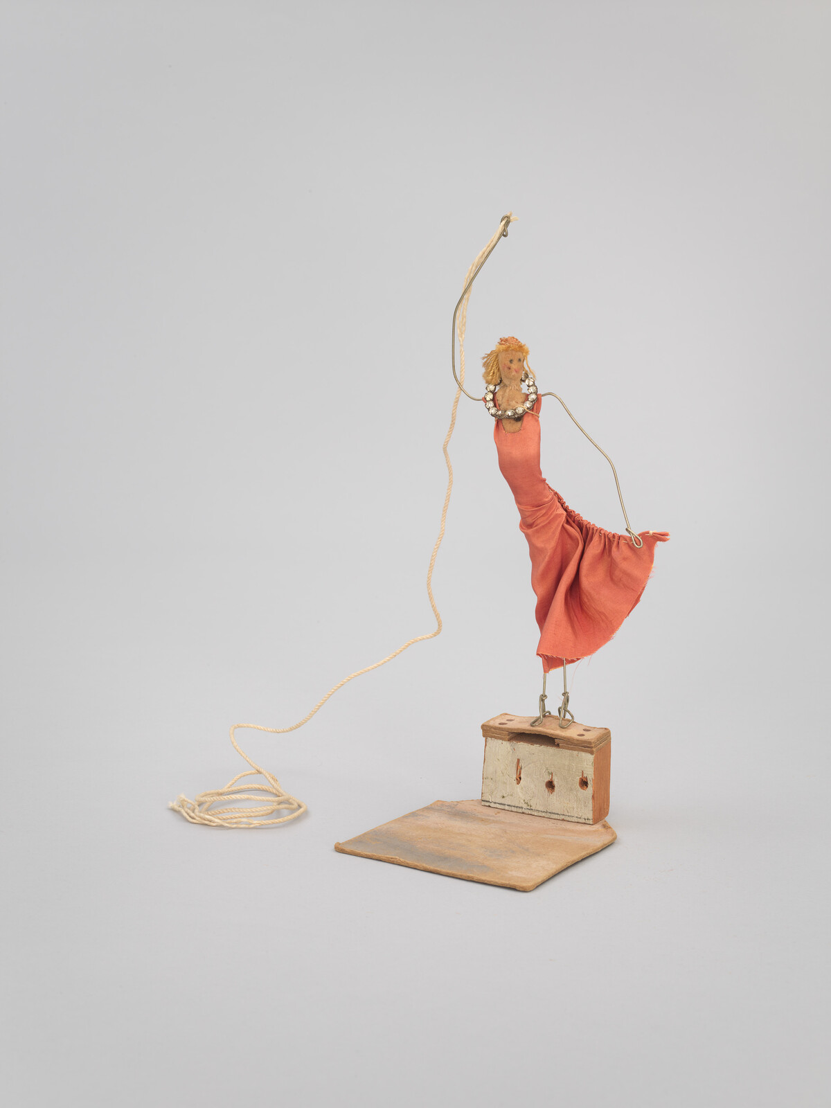 A small handmade wire puppet in a red dress balances on a wooden base while held by string.