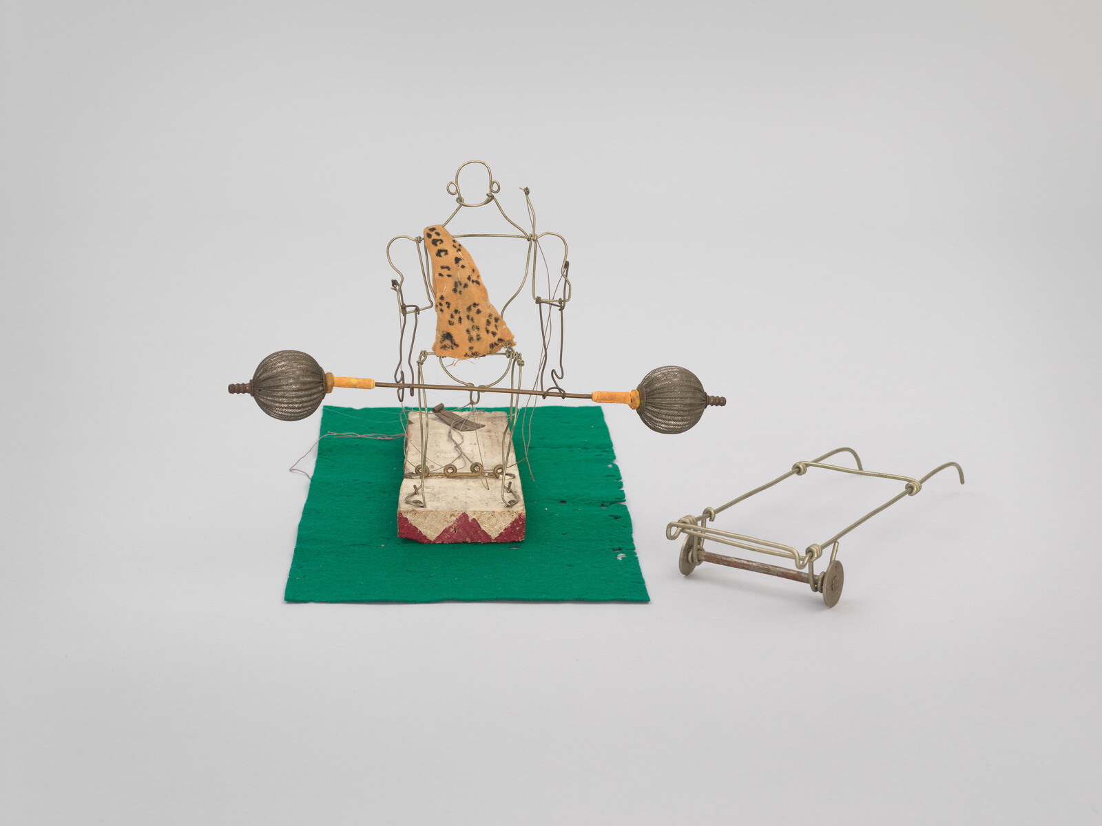A wire-framed figure seated on a block lifts a rod with two round weights on a green mat.
