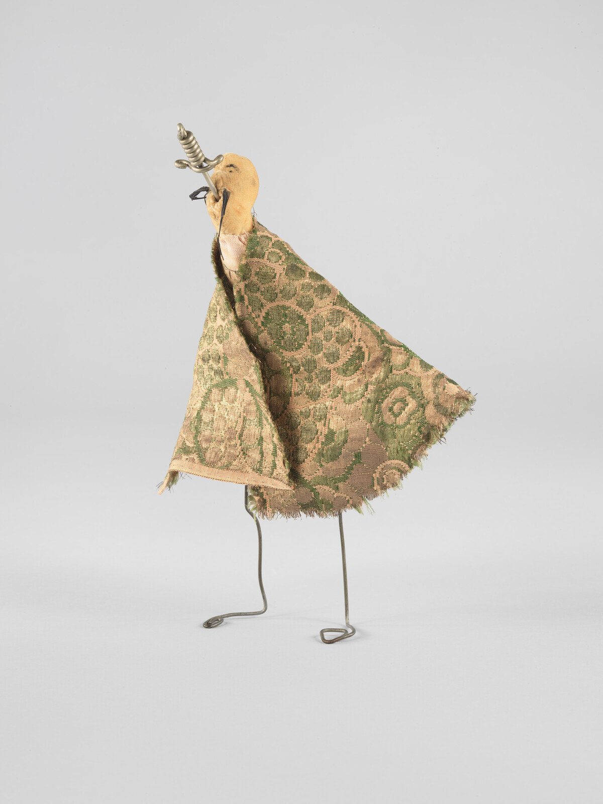 Small handmade puppet with patterned green cloak, wooden head, and bent wire legs standing upright.