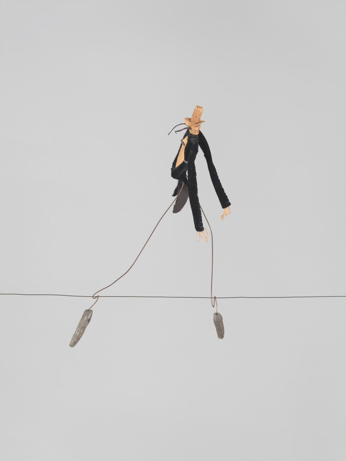 A handmade wire puppet wearing a tiny hat walks on a tightrope with small hanging weights.