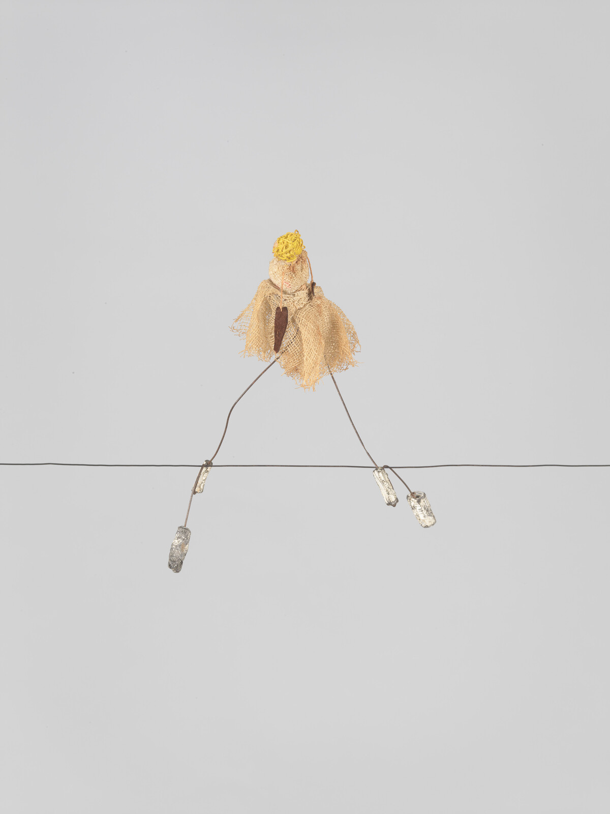 Tiny handmade doll with a yellow head and burlap skirt sits balanced on a thin wire.
