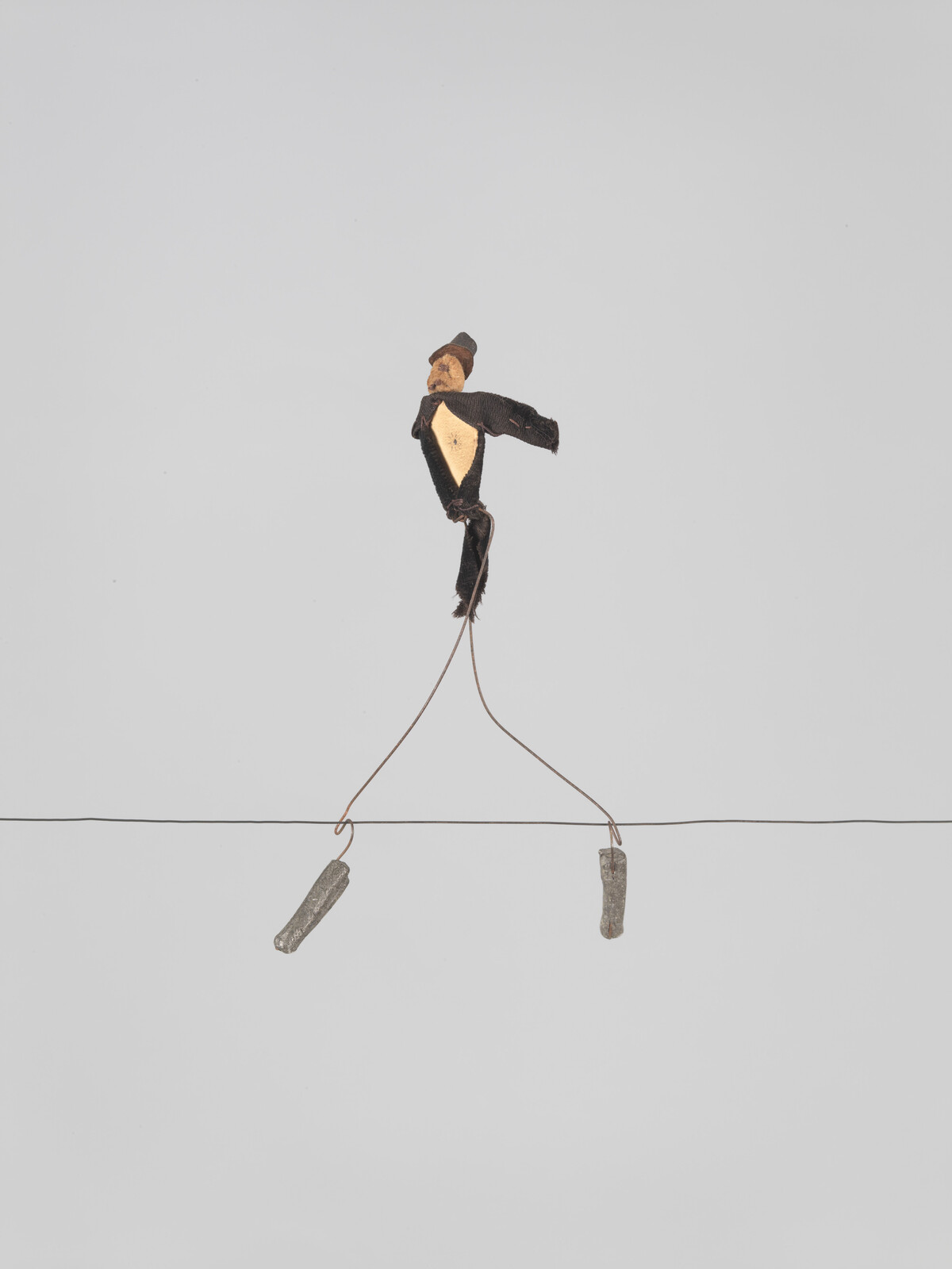 A small handmade doll balances on a thin wire with two weighted cords hanging below.
