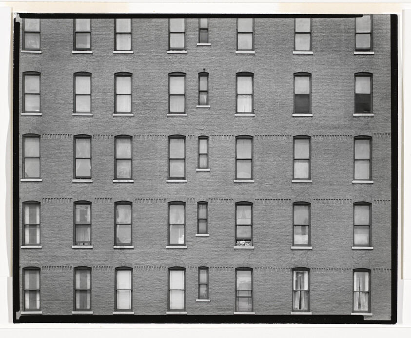 Brick apartment building facade with five rows of evenly spaced rectangular windows, some with curtains.