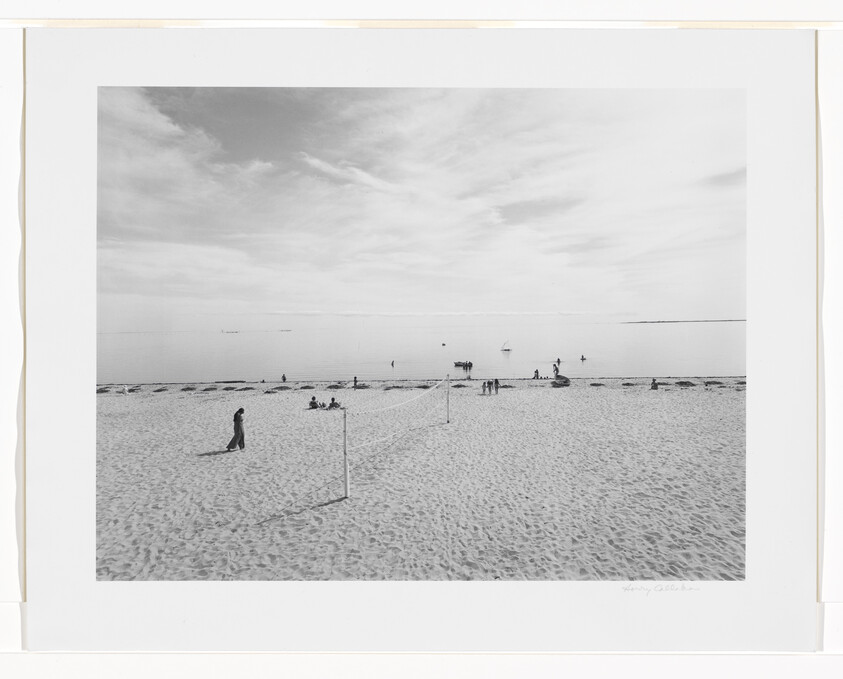 A quiet sandy beach with a volleyball net, a lone walker, and people near calm water.