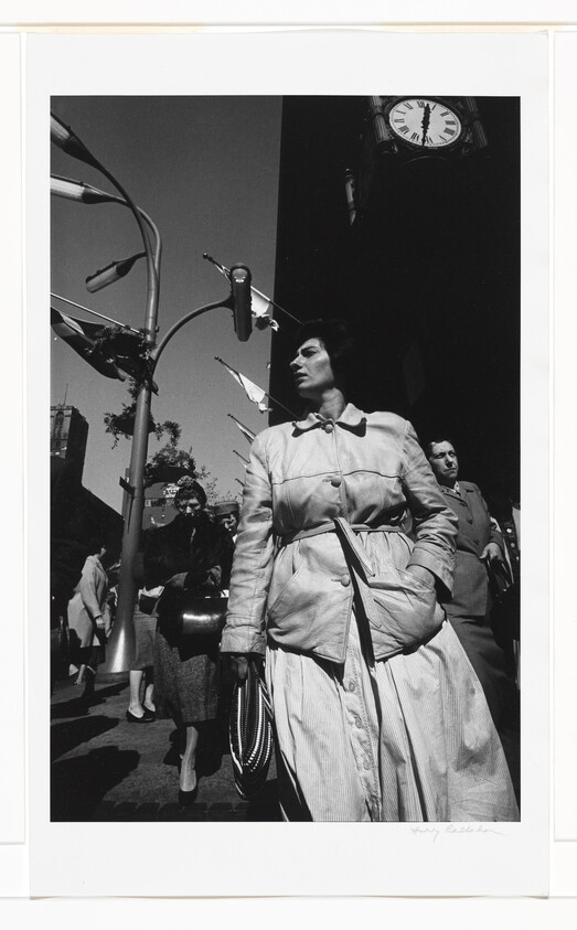 Woman in a belted coat walks through a city street with a large clock overhead.