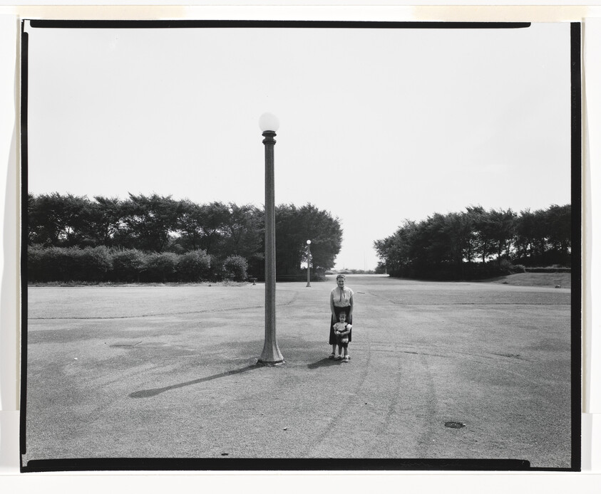 A woman stands with a small child beside a tall lamppost in a wide empty plaza.