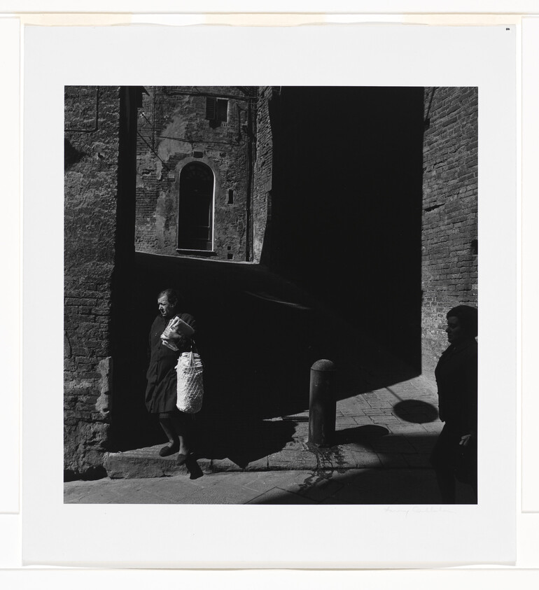 An older woman carries a large netted bag while walking through a sunlit street shadow.