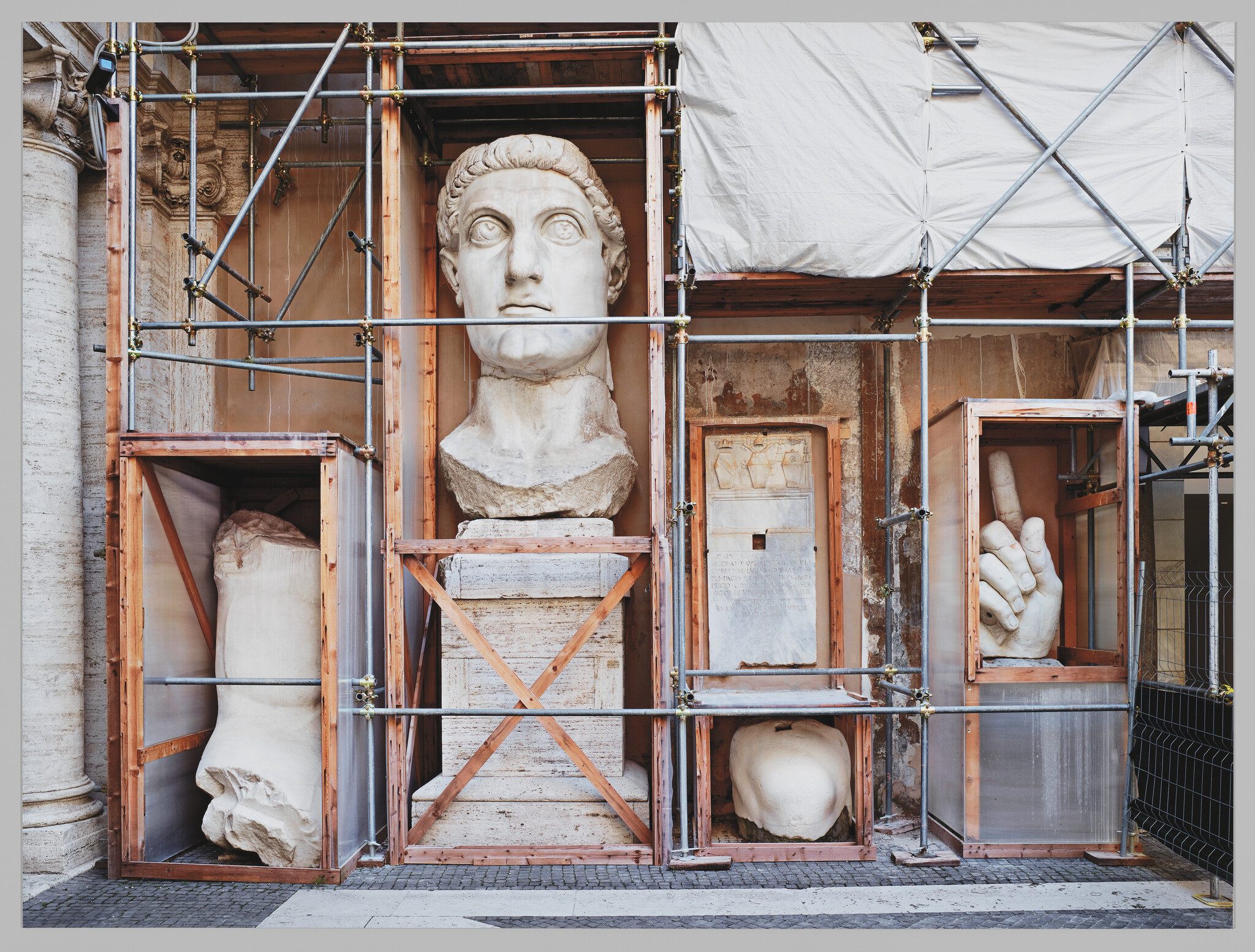 Large ancient marble statue fragments and a giant head are stored in wooden crates under scaffolding.