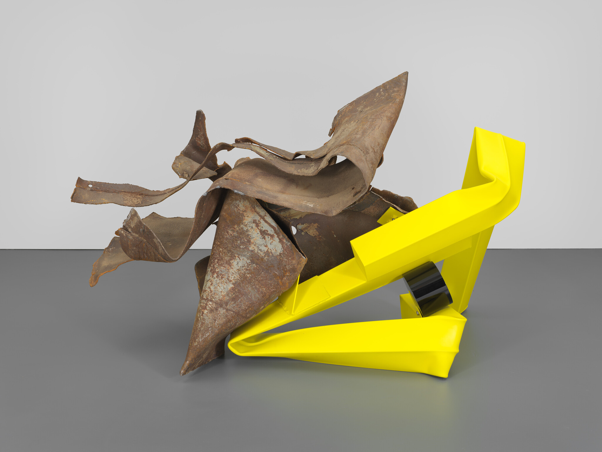Rusted twisted metal sheet entwined with a bright yellow folded geometric sculpture on gray floor.