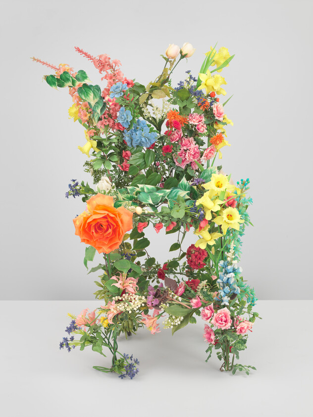 Chair-shaped sculpture covered in colorful flowers and green foliage standing on a gray surface.