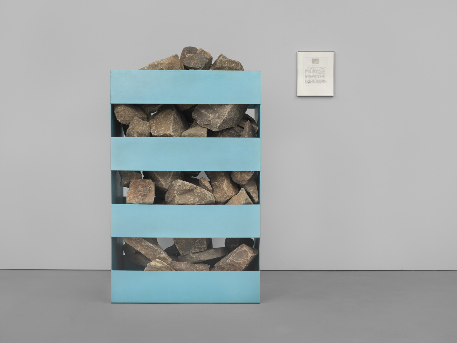 A tall blue shelf holds rough brown rocks stacked in four compartments against a gray wall.