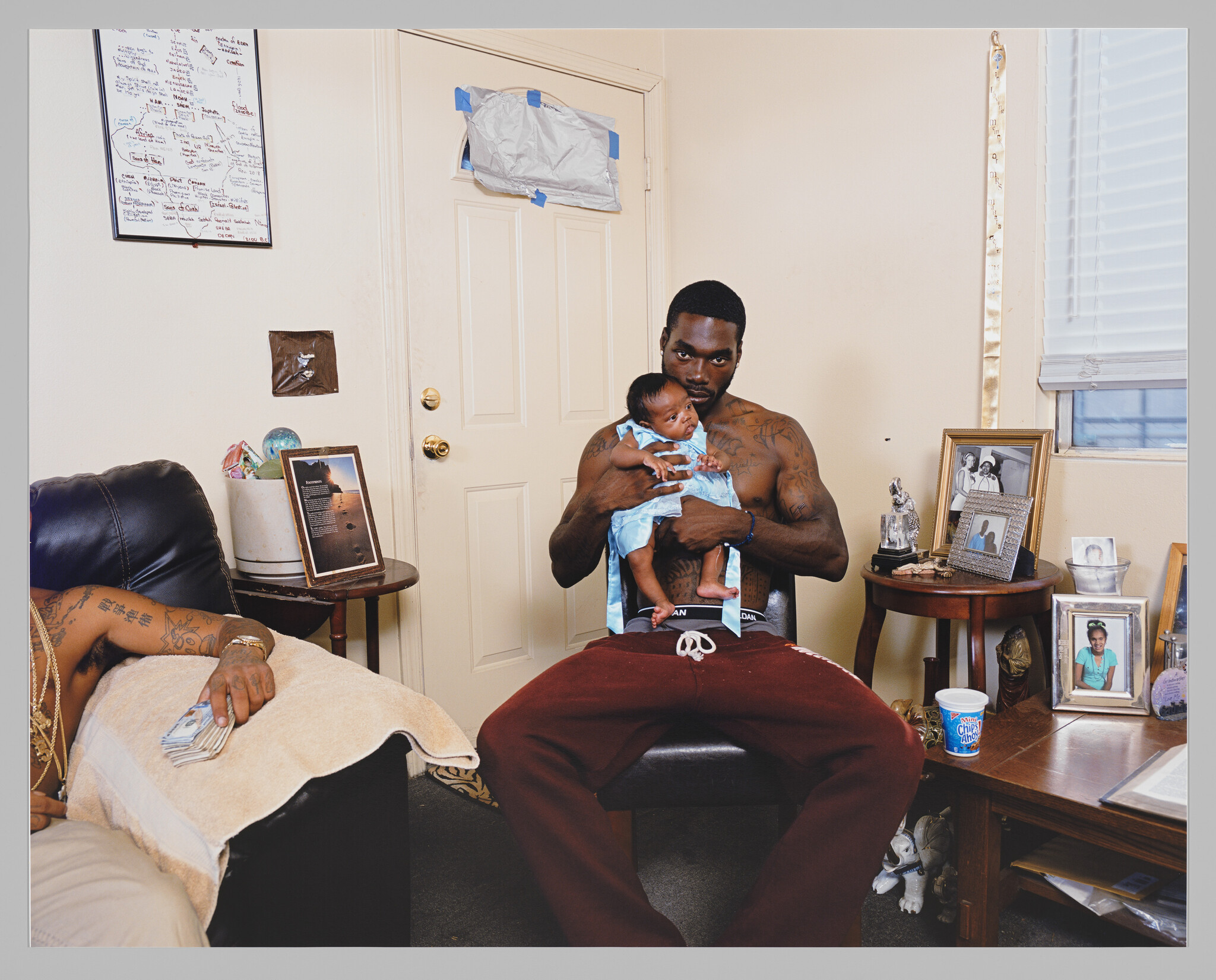 A shirtless man with tattoos sits in a chair holding a baby, surrounded by personal items and photographs in a room with a closed door and a window.