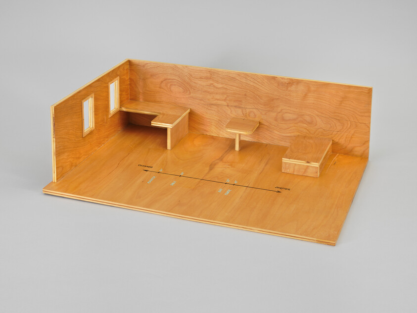 A small wooden room model with two windows, a wall desk, a pedestal table, and floor measurement markings.