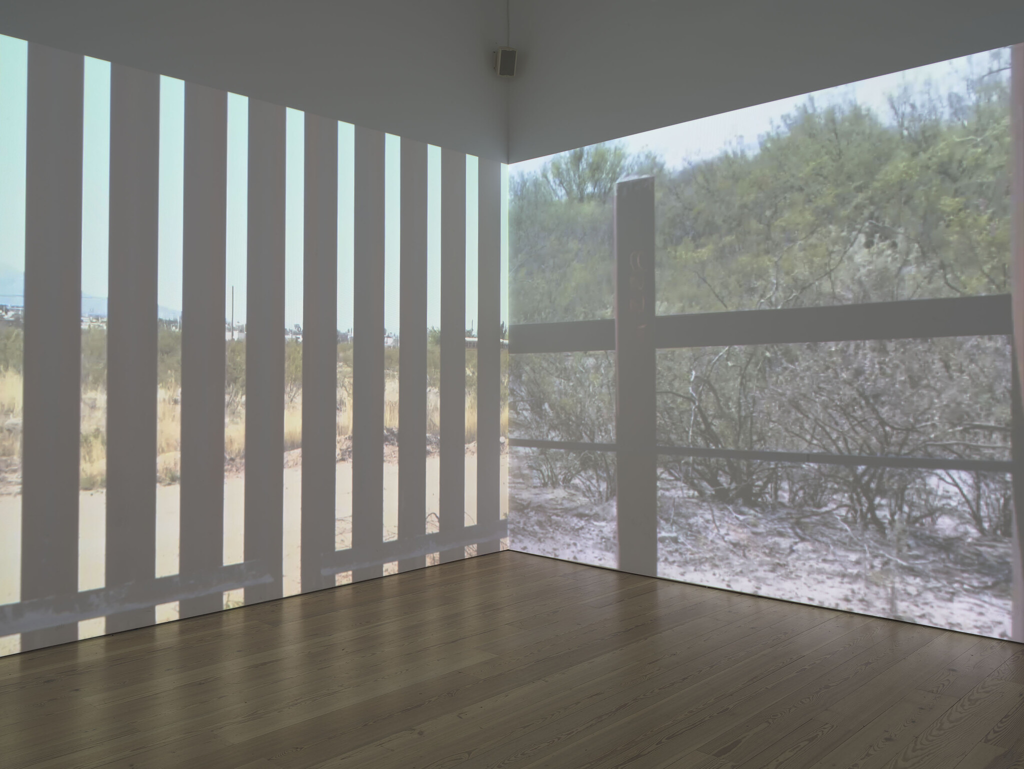 A gallery room with projections of a desert landscape and vertical fence shadows across the walls.