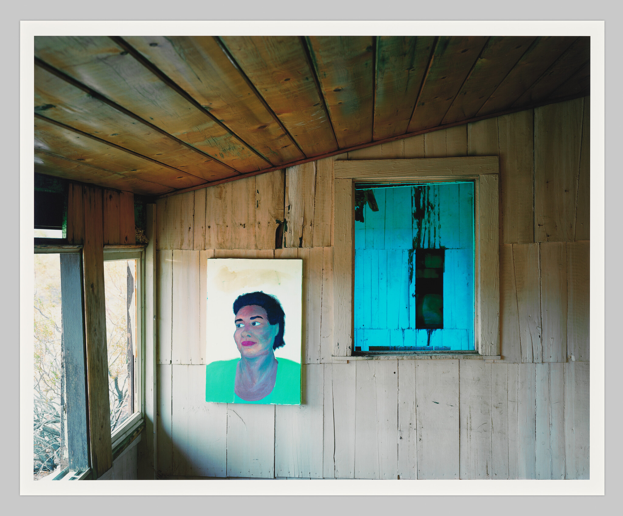 A painted portrait of a woman hangs on a weathered wooden wall beside a bright blue window.