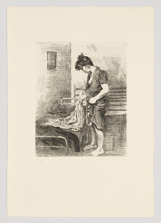 A barefoot young woman stands beside a bed folding or inspecting a sheet in a simple room.