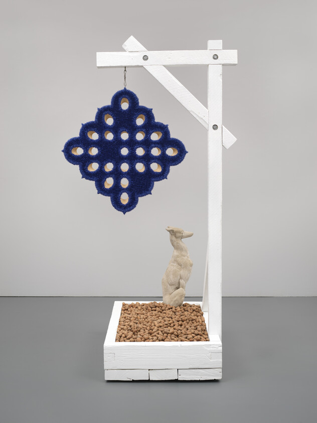 A small animal sculpture sits on pebbles under a white wooden frame with a blue patterned object hanging.