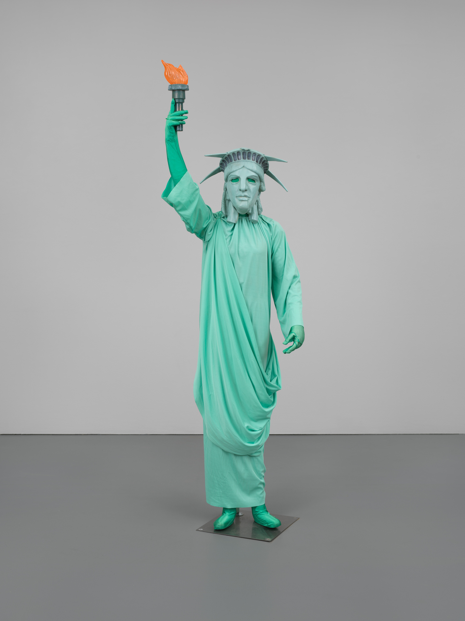 Person dressed as the Statue of Liberty, holding a torch aloft, standing against a plain gray background.