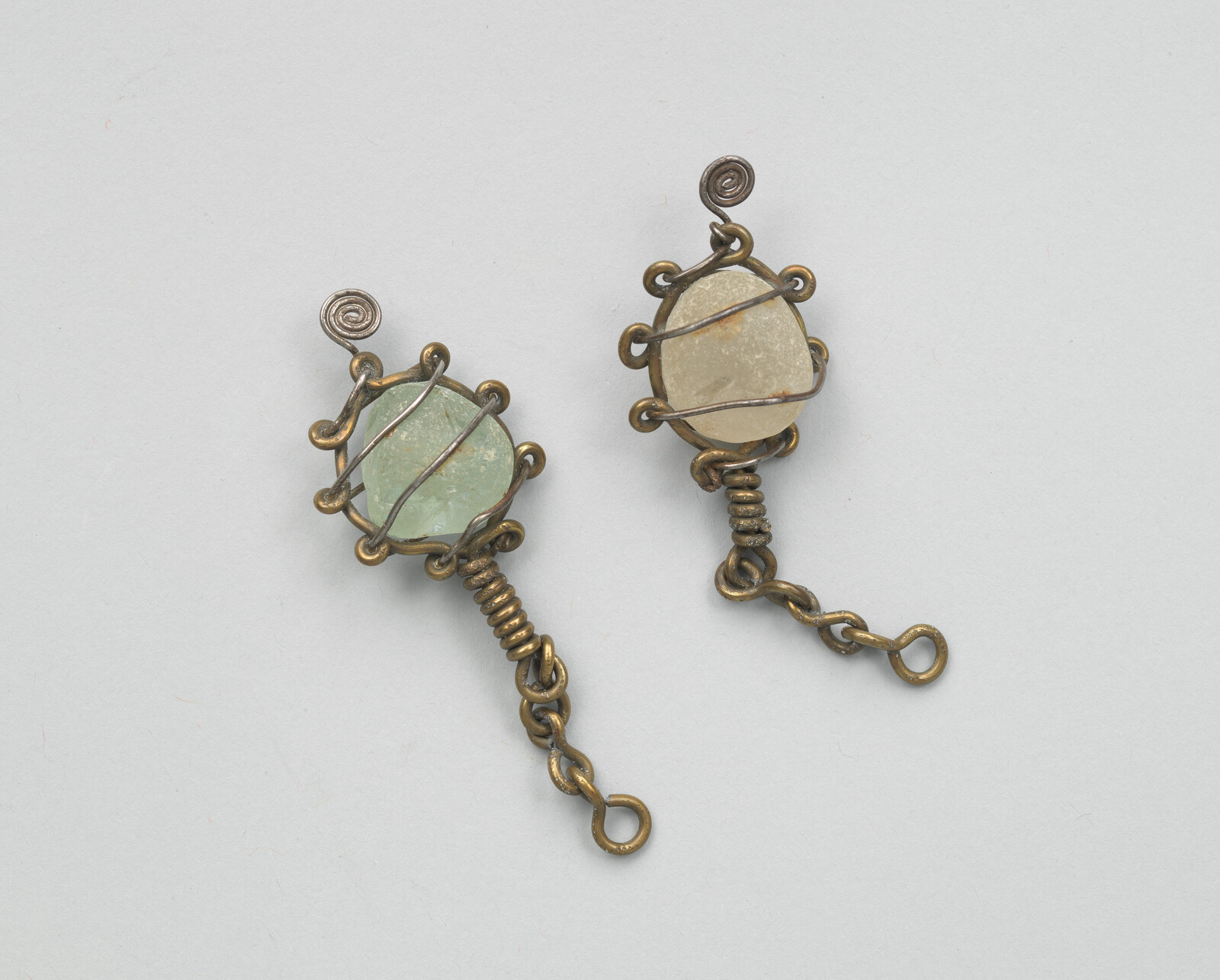 Two small brass wire-wrapped pendants each holding a rough pale green or beige sea glass stone.