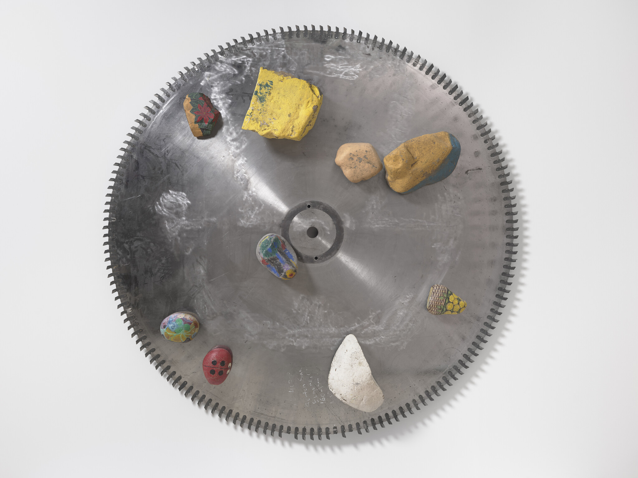 Large circular saw blade mounted on wall with several small painted rocks arranged on its surface.