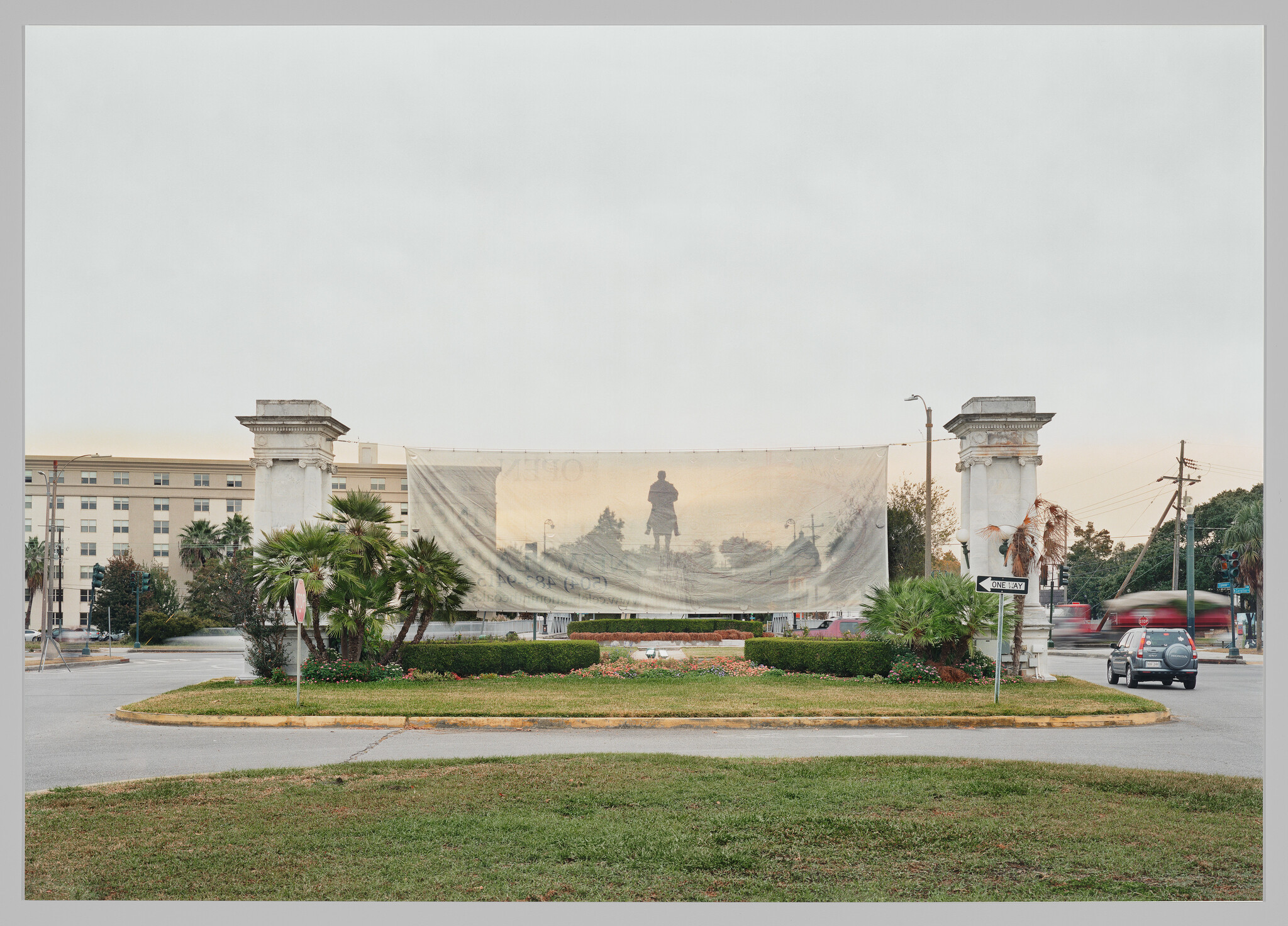 A monument of a man riding a horse behind a translucent banner hung between two columns on a grassy street island