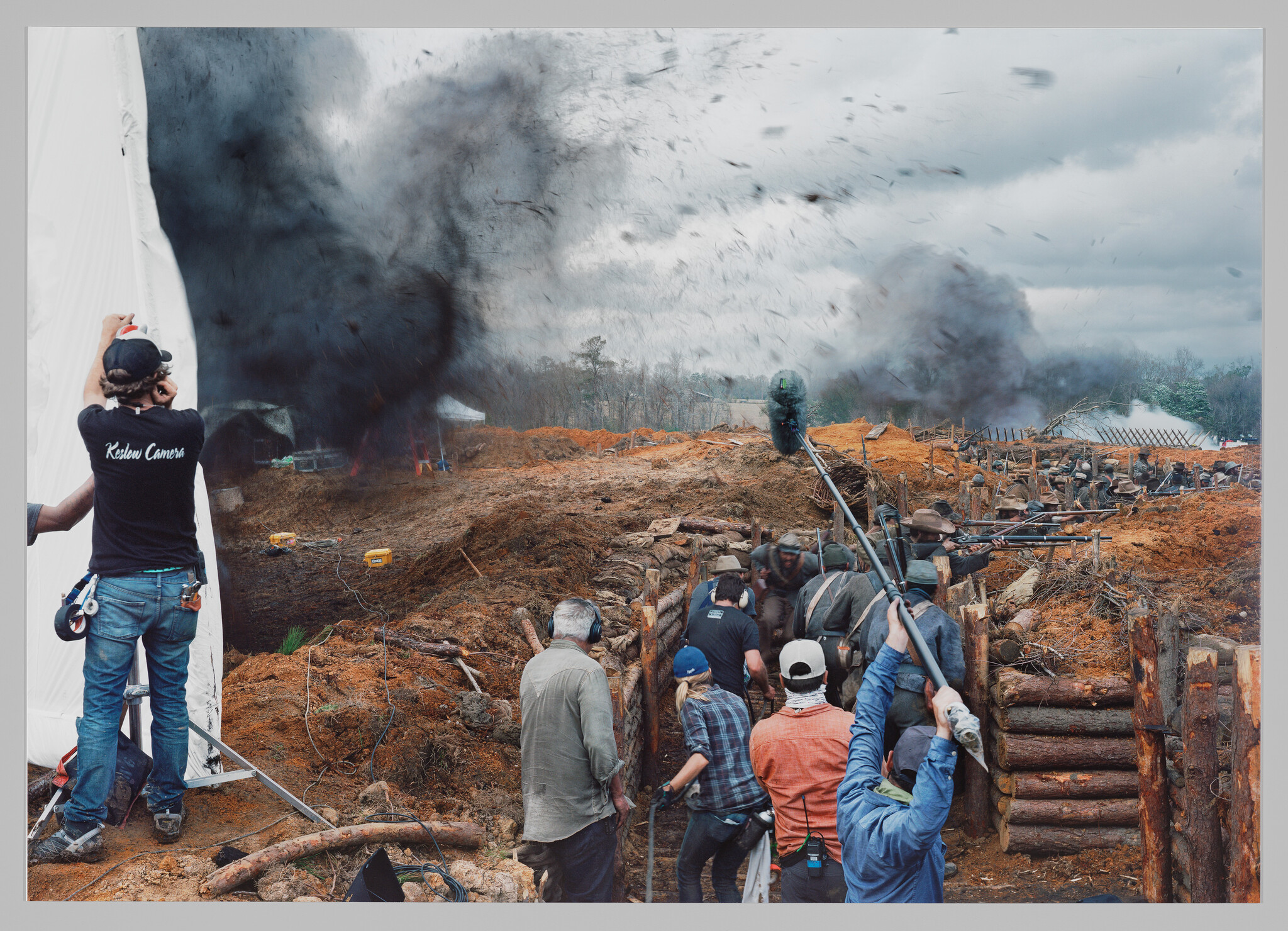 Film crew captures a battle scene with actors in trenches, surrounded by smoke and debris. Crew members hold boom mics and cameras.