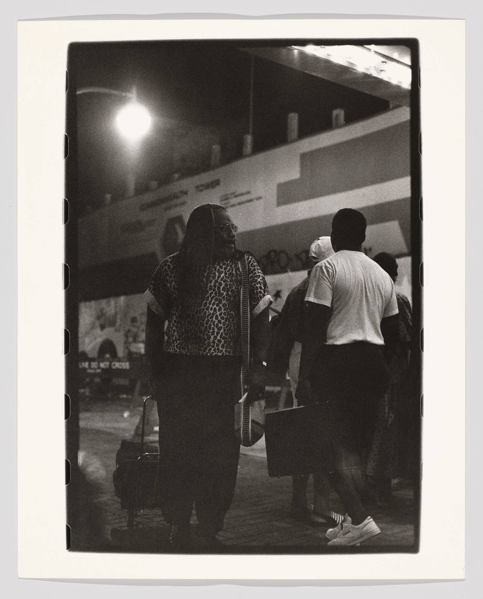 A woman in a leopard-print top pulls a rolling bag while people walk past under a streetlight.