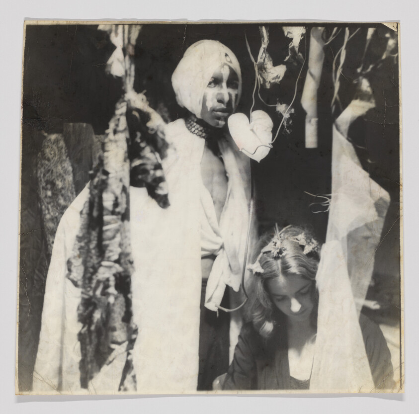 Man in a turban and robe stands behind a seated woman wearing a floral headpiece, heart ornament hanging.