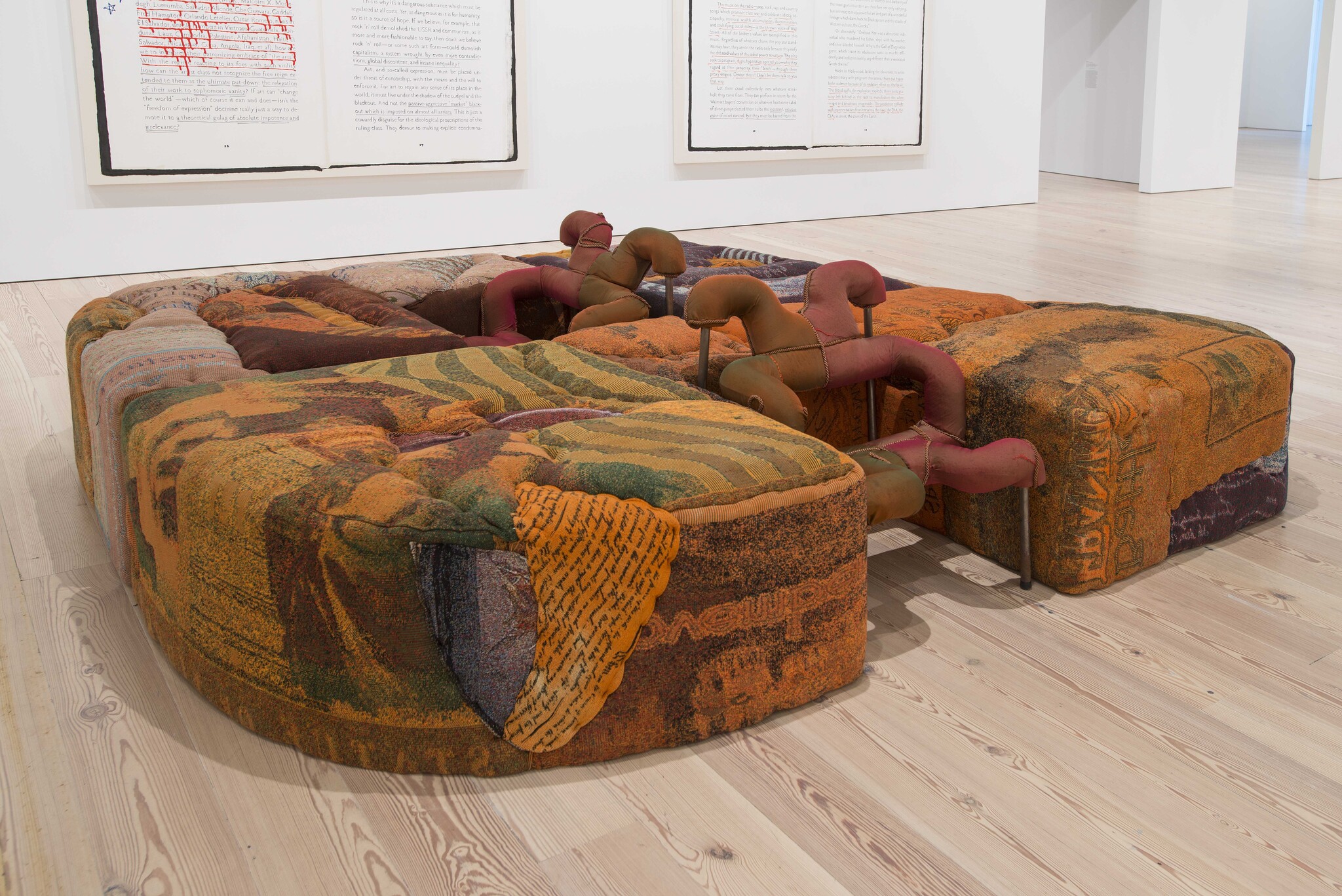 Large, colorful, fabric-covered sculpture with soft, rounded shapes and pipe-like forms, displayed in a modern gallery.