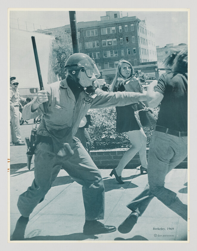 Police officer in helmet swings a baton while grabbing a protester on a city sidewalk.