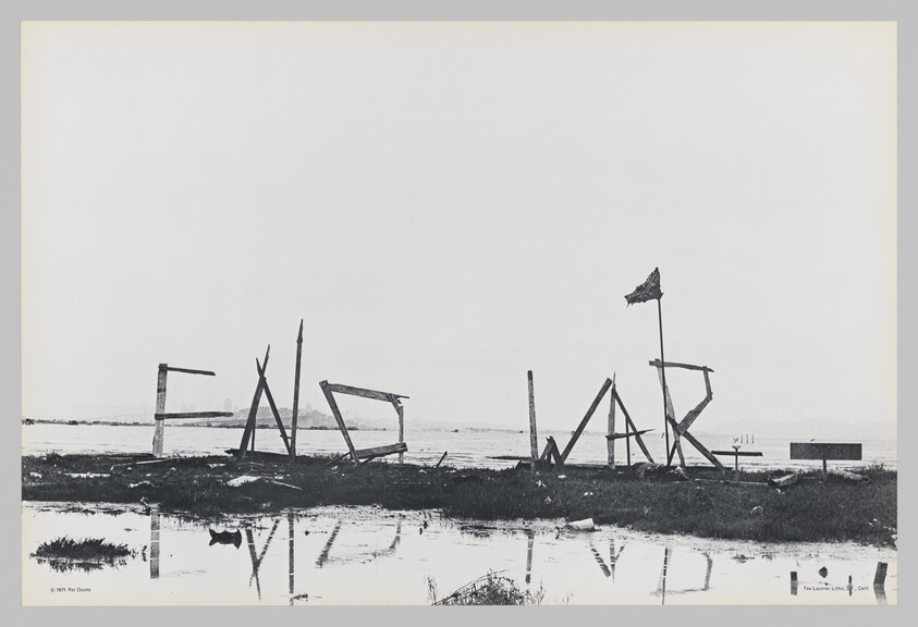 Large makeshift wooden letters spell out a broken sign along a flooded shoreline, reflected in water.
