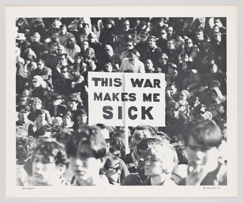 A black and white photograph capturing a dense crowd of people at a protest with a prominent sign held up that reads "THIS WAR MAKES ME SICK." The faces of the individuals are mostly indistinct, conveying the collective nature of the event. The photo is credited to Janos Panitz and Steve Sachs, 1987.